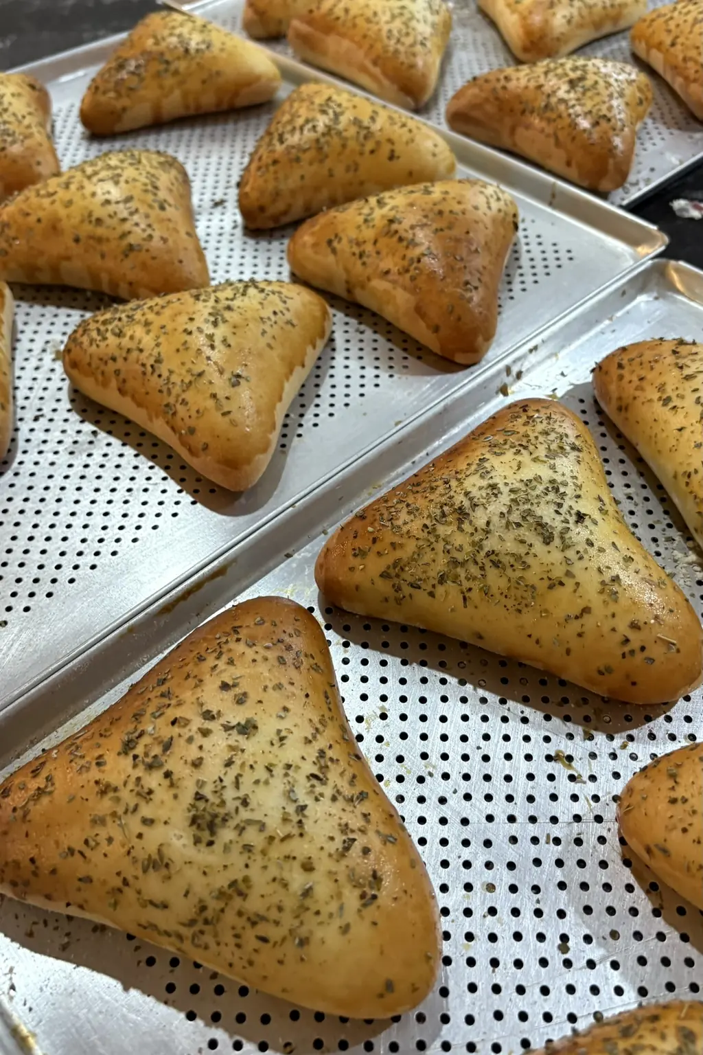 Foto de celular: esfihas assadas em assadeiras de alumínio furadas, na cozinha.